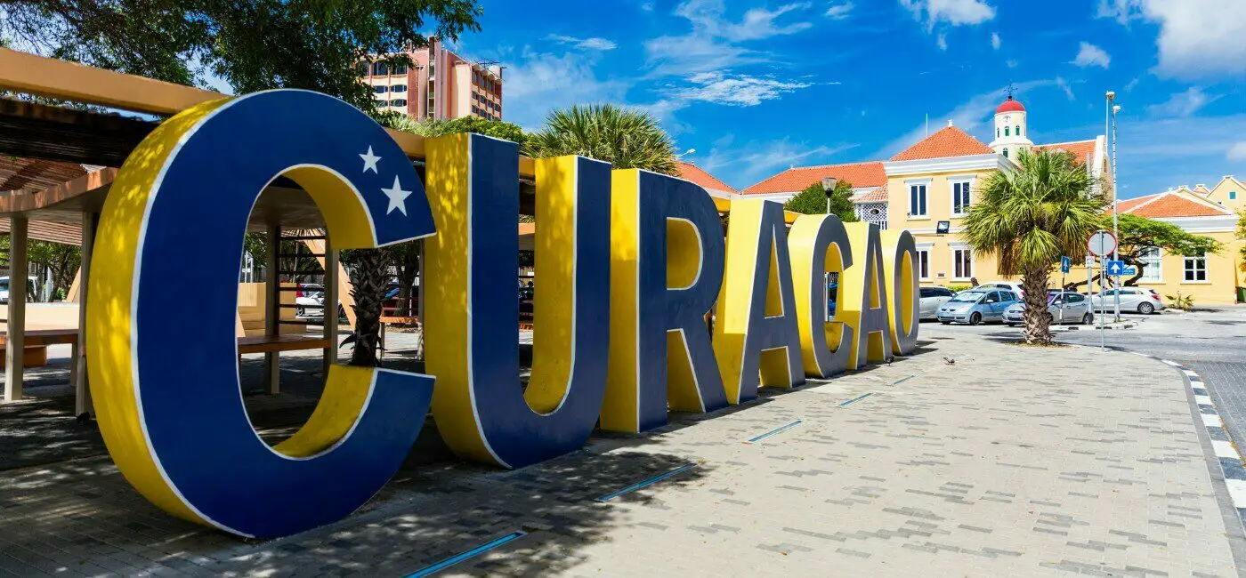 Large yellow and blue CURAÇAO sign in Willemstad with colonial buildings and palm trees in the background